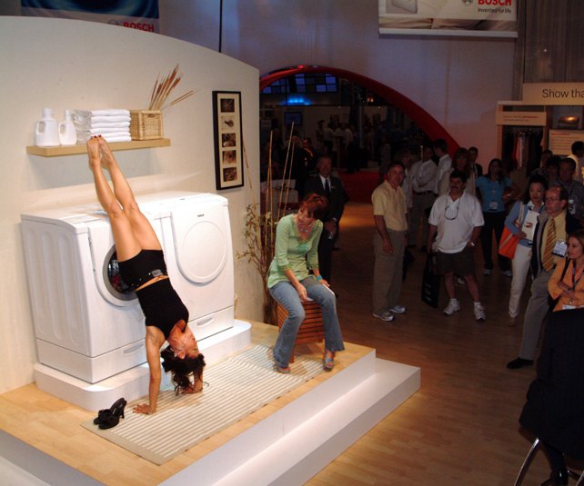 A performer doing a handstand on a mat at a laundry-appliance booth, her feet raised into the open door of a washing machine, while a seated assistant watches and spectators look on.