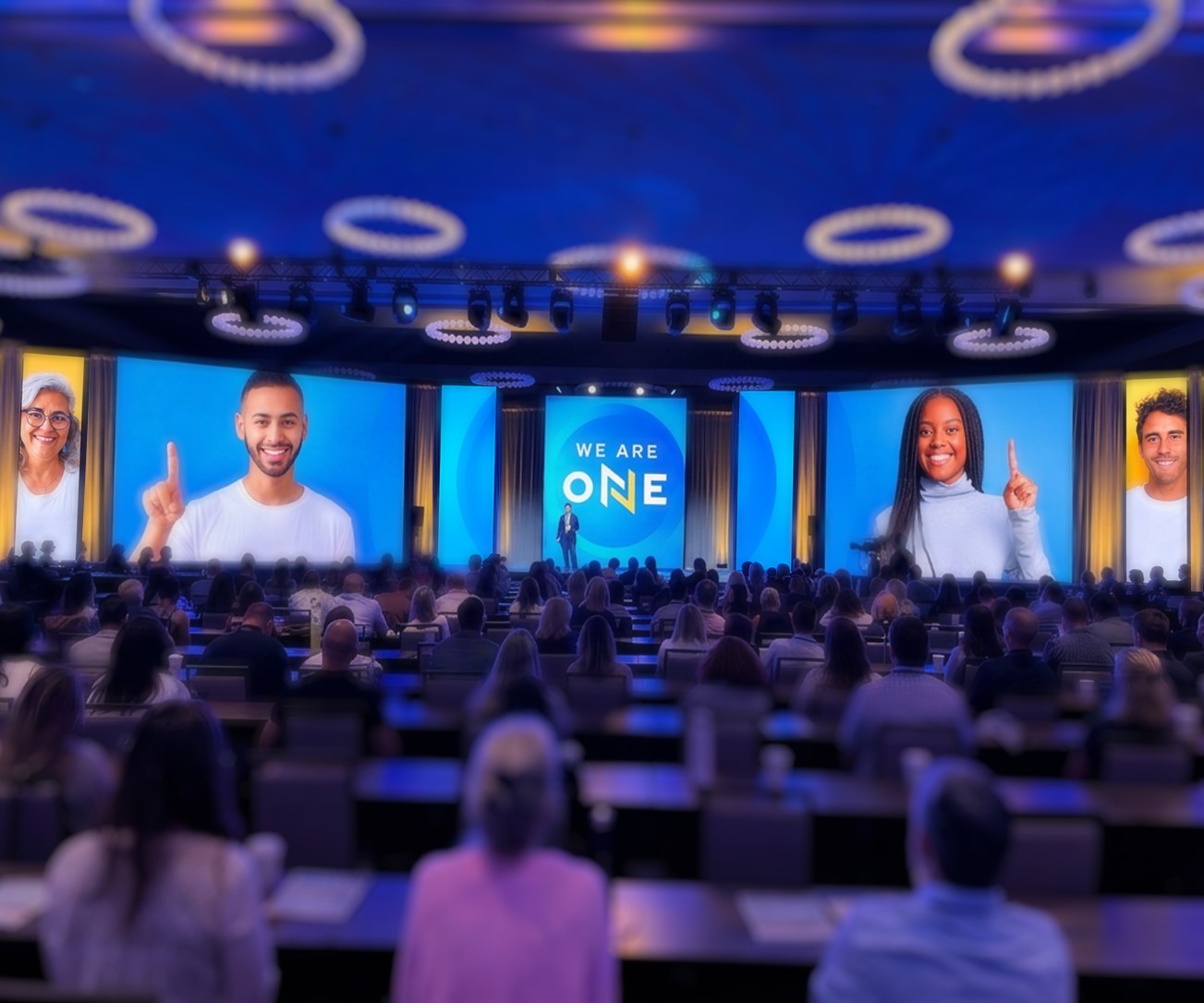 A nine-screen general session stage: large blue video panels with employee portraits flank a centre screen reading We Are One, above a seated audience.