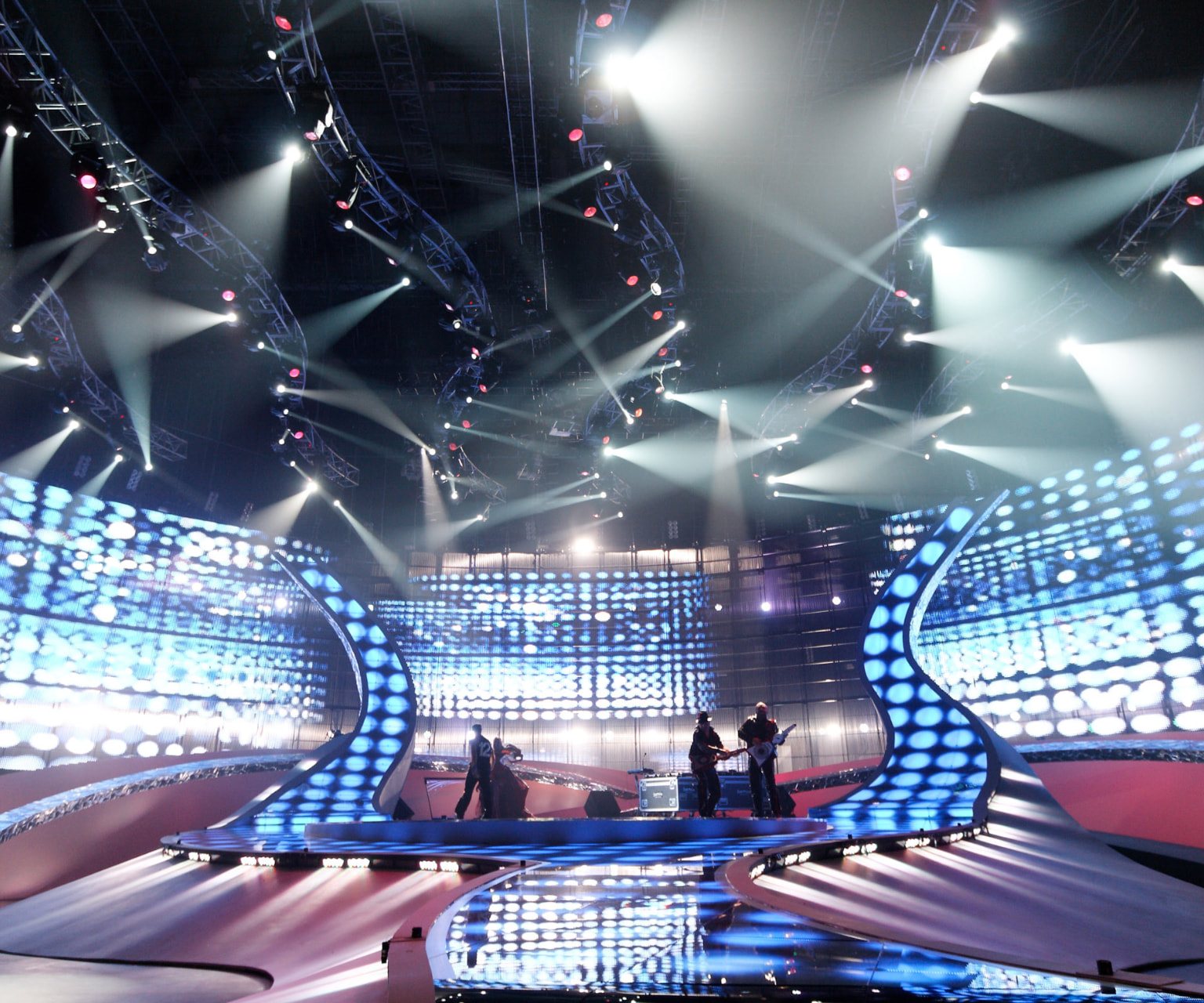A stage lit by curved glowing arches and overhead beams of light, with a large curved LED backdrop and silhouetted performers on the central platform.