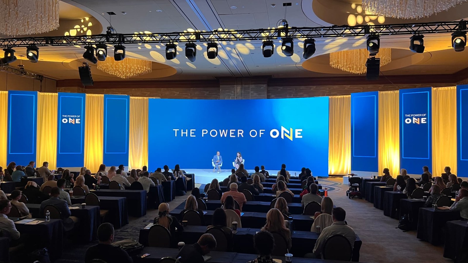 A blue and yellow conference stage displaying The Power of One, viewed from the audience.