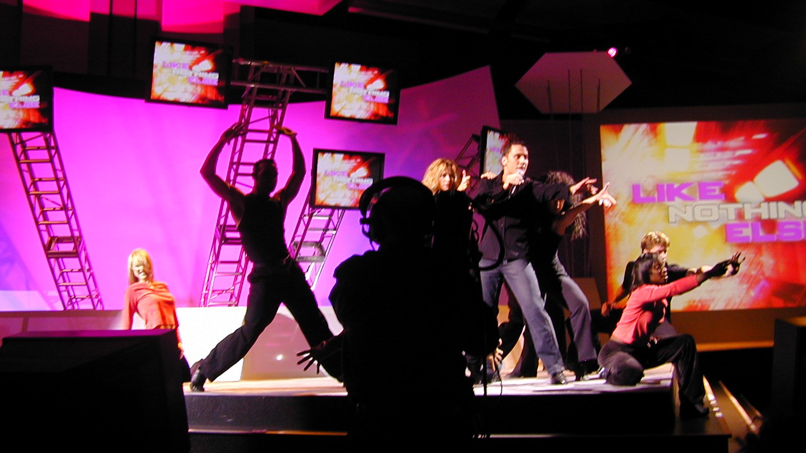 Performers in motion on a vivid pink stage, with a camera operator in the foreground.