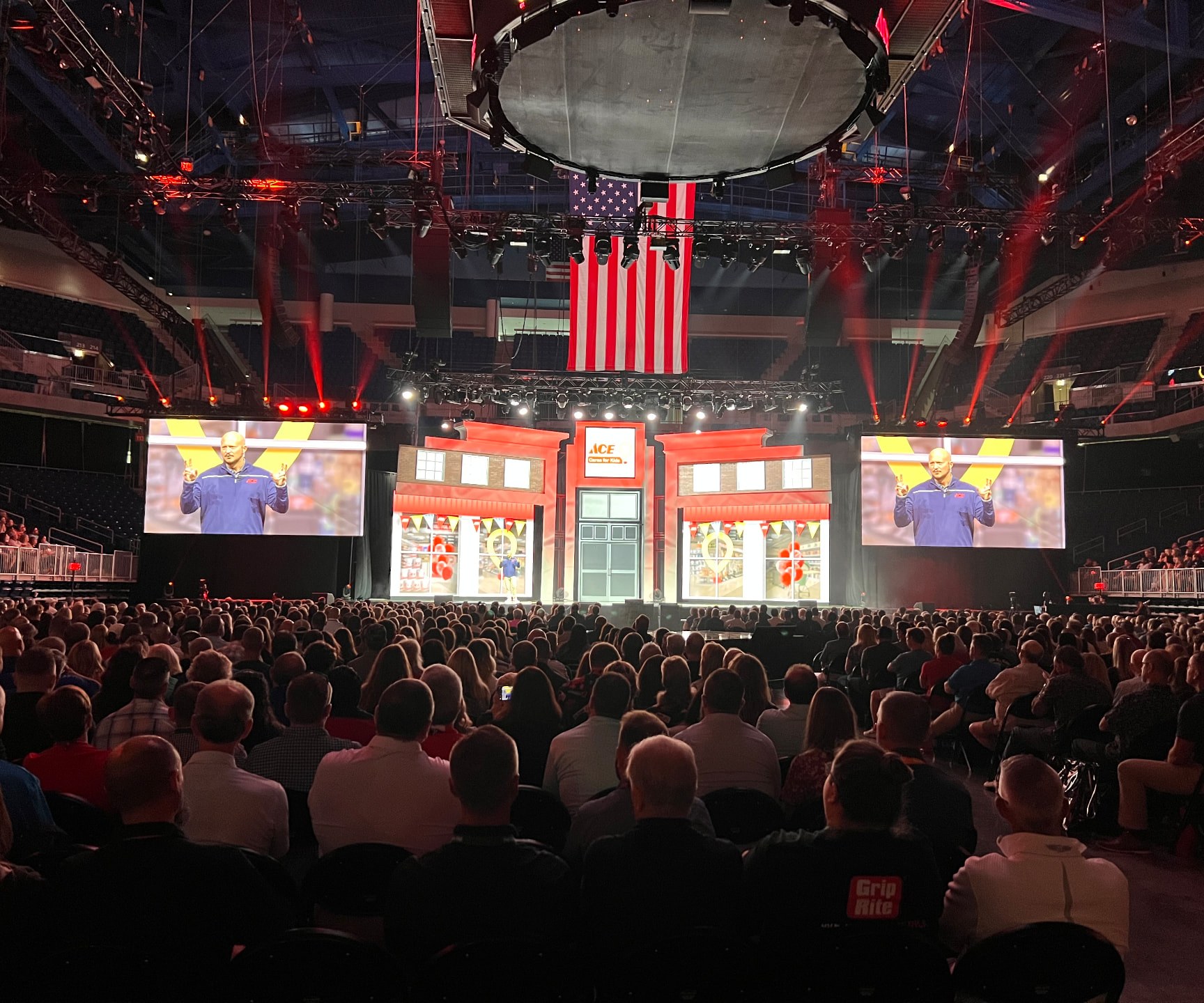 A speaker on an arena stage built like an Ace storefront, flanked by two large video screens, with a seated audience in the foreground.